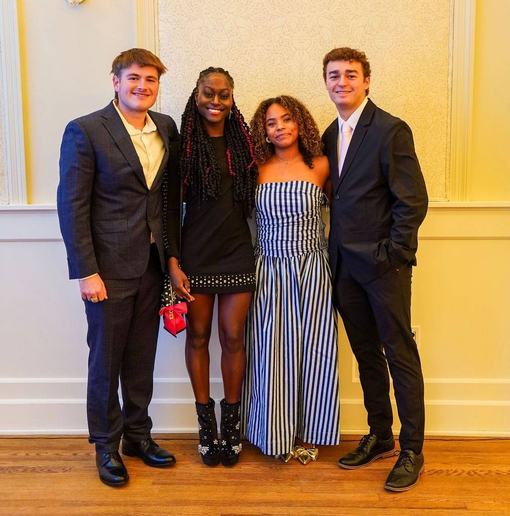 Four people posing together indoors, dressed formally, against a cream wall.