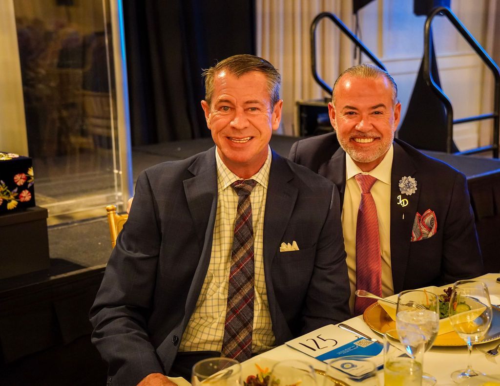 Two men in suits seated at a restaurant table, smiling near wine glasses and a plated meal.