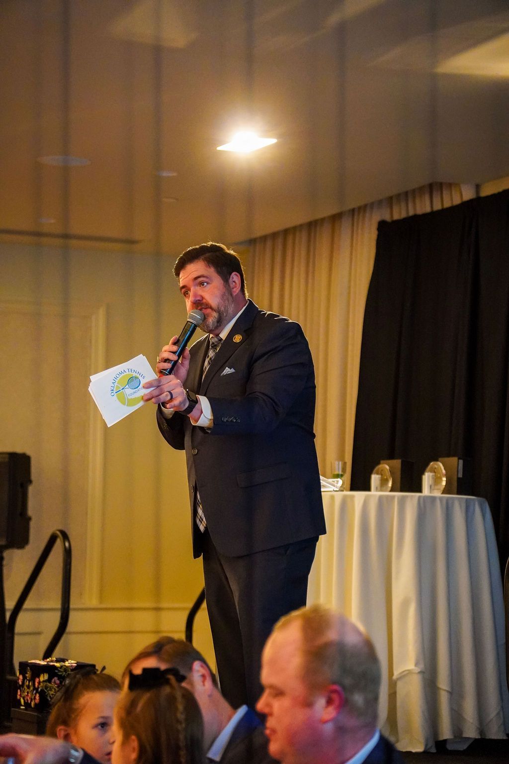 Man speaking into a microphone onstage, holding notes, with a lit table and curtains behind him.