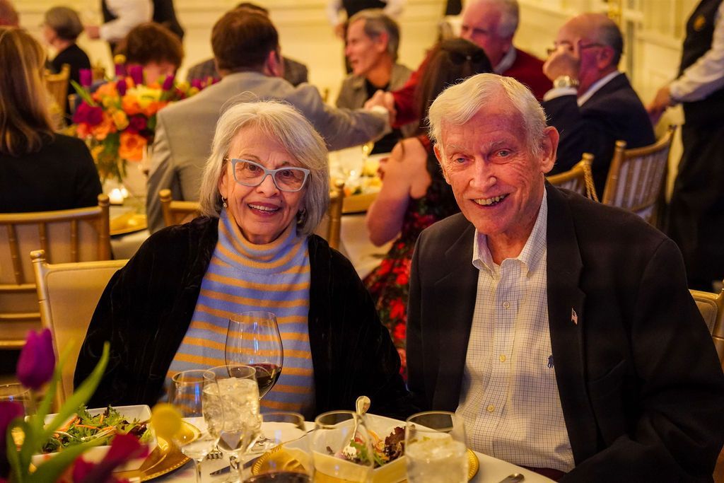 Two smiling people seated at a banquet table with flowers and glassware in a warmly lit hall