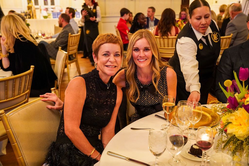 Two women smiling at a banquet table with wine glasses and flowers in a warm-lit dining hall