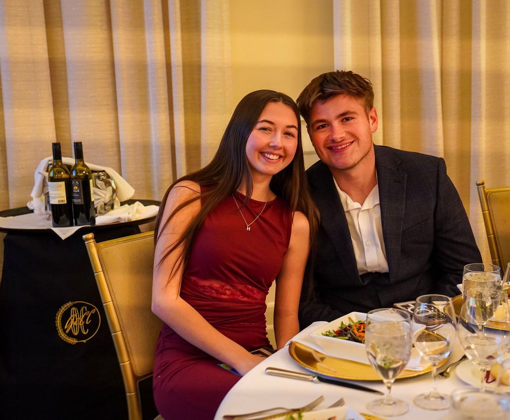 Smiling couple seated at a formal dinner table with wine glasses and plated food, in a warmly lit restaurant