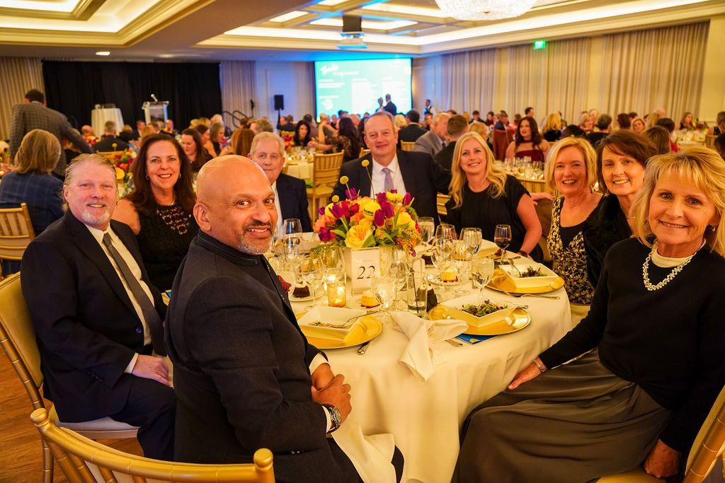 People seated around a banquet table at a formal event, smiling in a brightly lit ballroom.