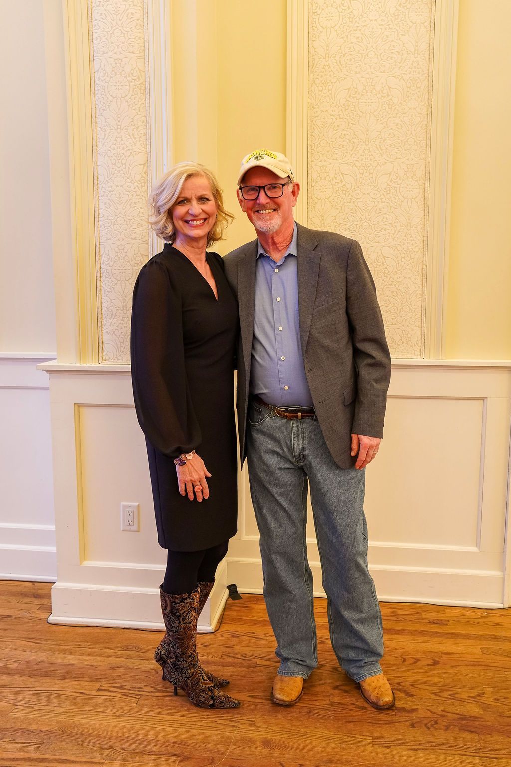 Two people standing and smiling in a warmly lit room with cream walls and wood flooring.