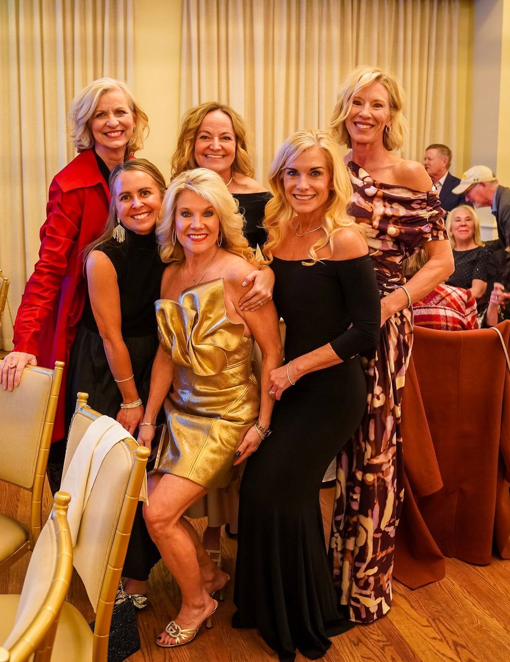 Five women posing at a formal event, wearing elegant dresses and smiling in a banquet hall.