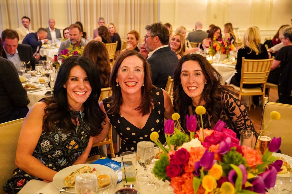 Three women smiling at a banquet table with colorful flowers and candles, in a warmly lit event hall