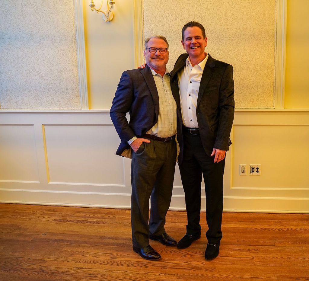 Two people in suits smiling and standing together in a warmly lit room with wood floors.