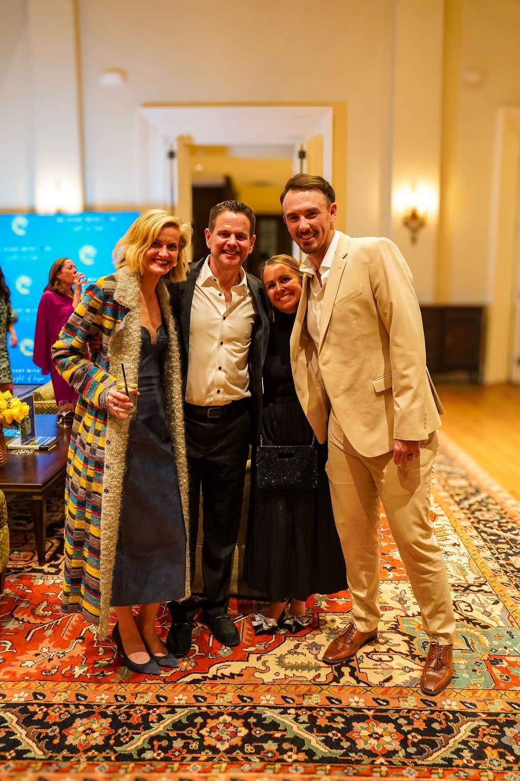 Four people posing together on a patterned rug in a warmly lit room, smiling at the camera.