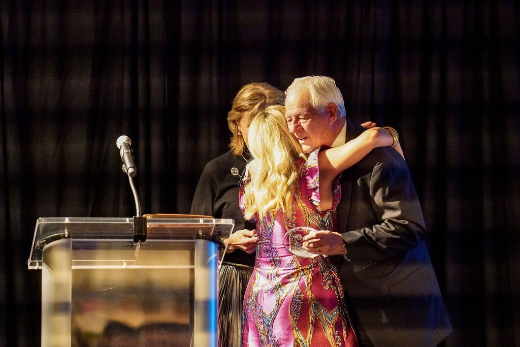 Two people embracing beside a podium and microphone on a stage, with dark curtains behind them.