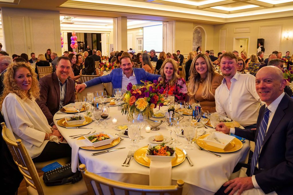 Group of people seated at a banquet table in a bright ballroom, smiling with flowers and plated food.