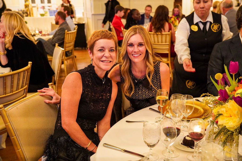 Two women smiling at a banquet table with wine glasses and a floral centerpiece.