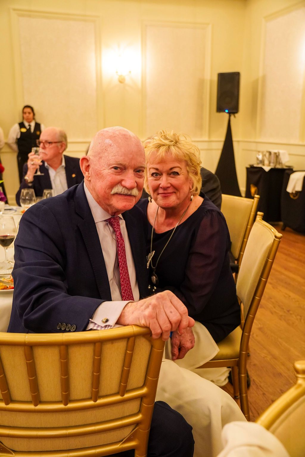 Two smiling people seated at a banquet table in a warmly lit ballroom
