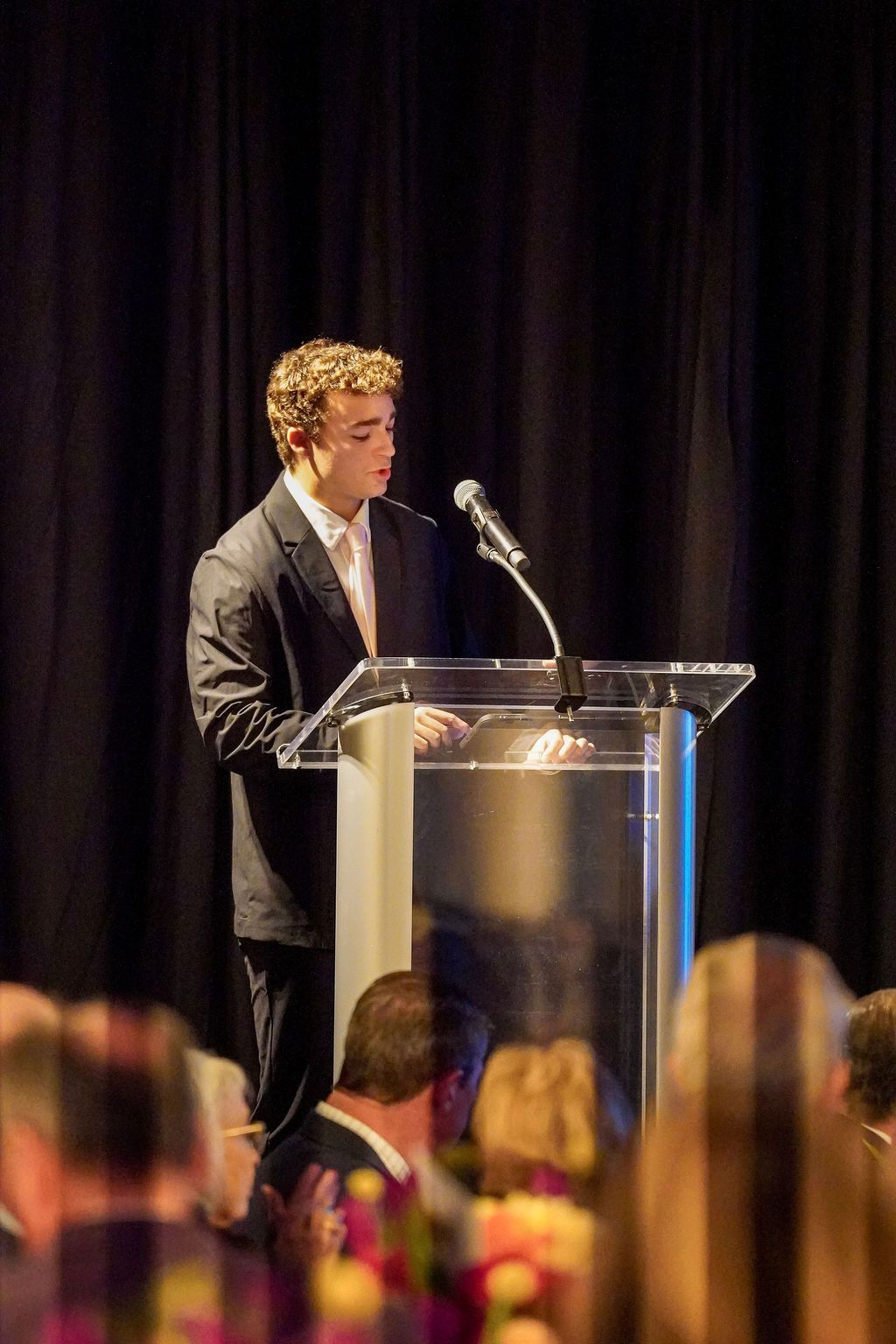 Man speaking at a podium with a microphone, facing an audience in a dimly lit venue