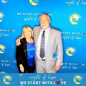 Two people posing in front of a blue “Night of Hope” event backdrop with “We Start With Love” text.