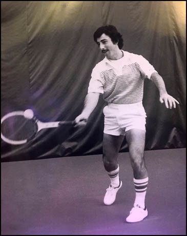 Black-and-white photo of a tennis player mid-swing on an indoor court, wearing striped shirt and white shorts.