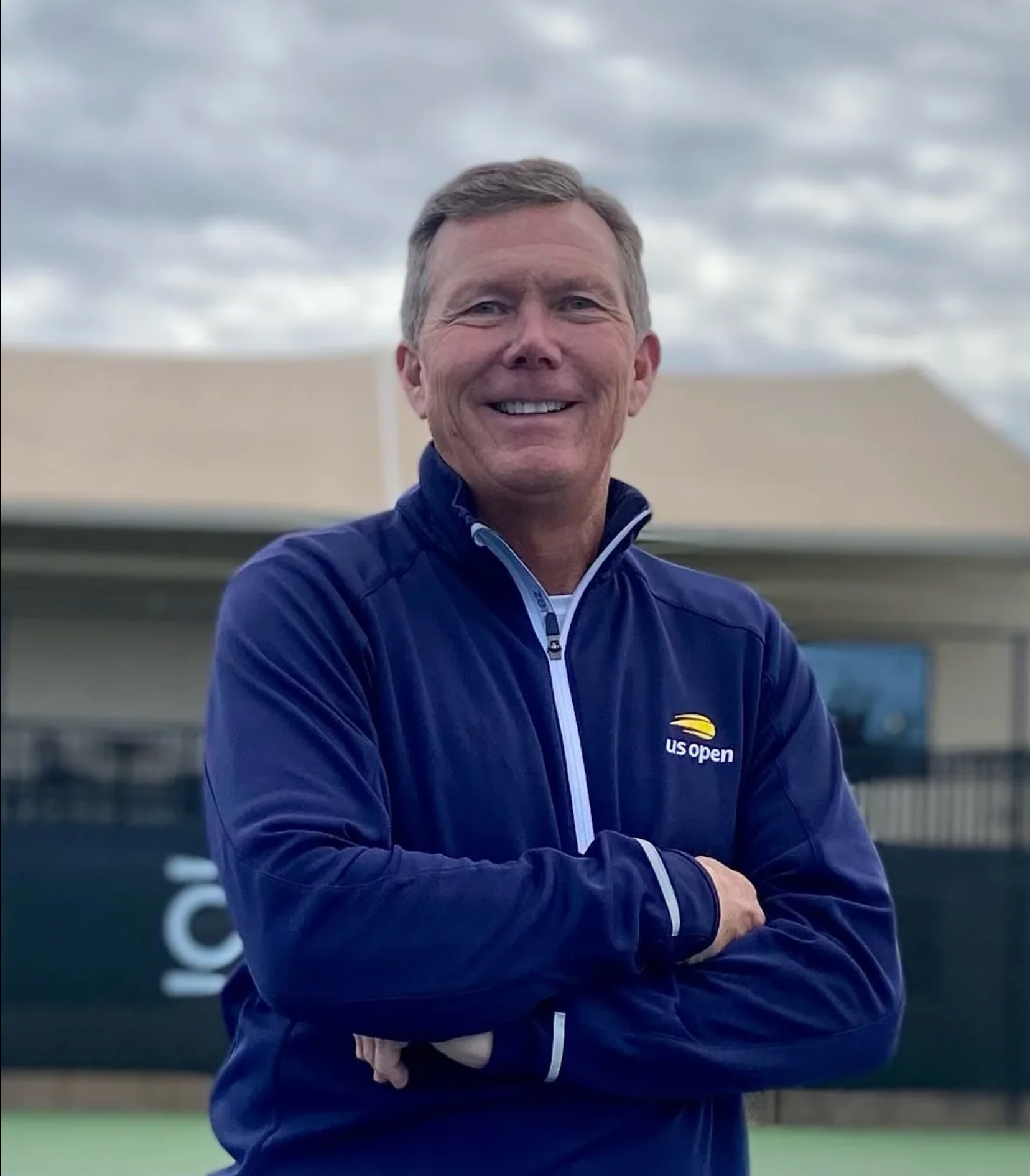 Smiling man in a blue jacket with arms crossed outdoors in front of a building