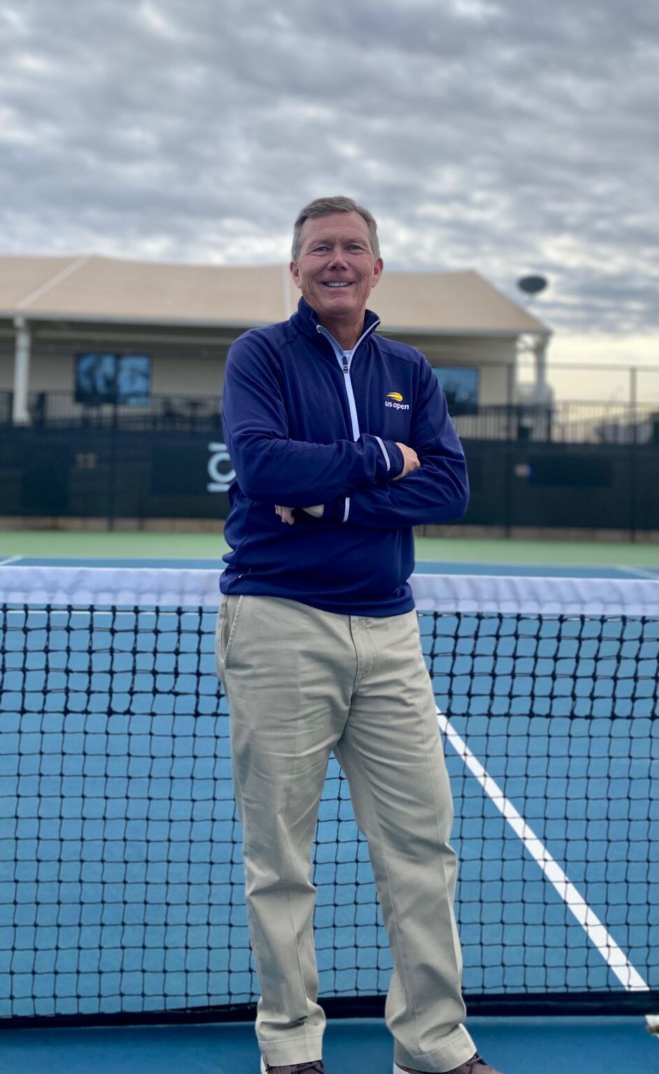 Man standing with arms crossed on a tennis court beside a blue net, wearing a navy sweater and khaki pants.