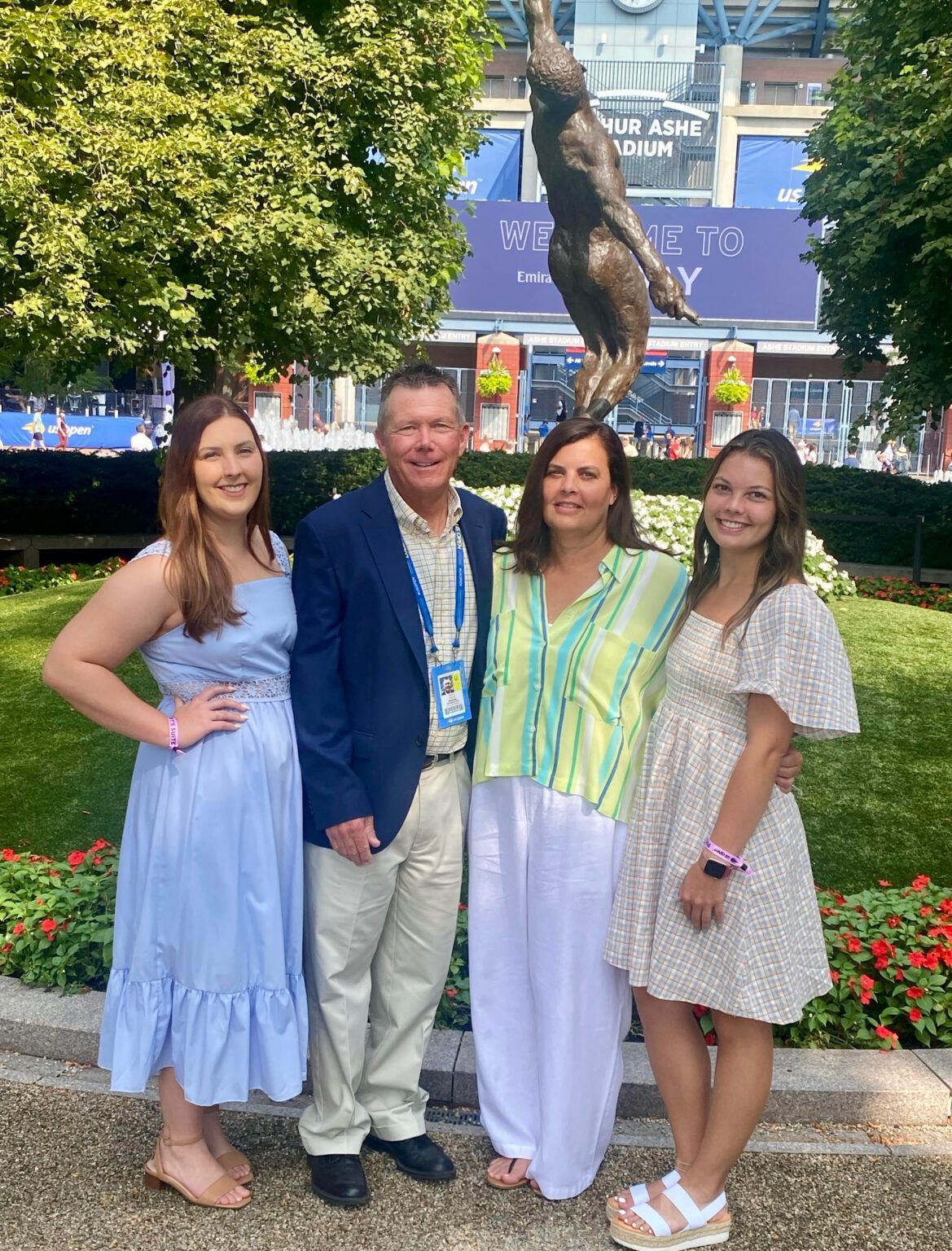 Four people posing outdoors in front of flowers and a fountain, with a building and banner behind them.