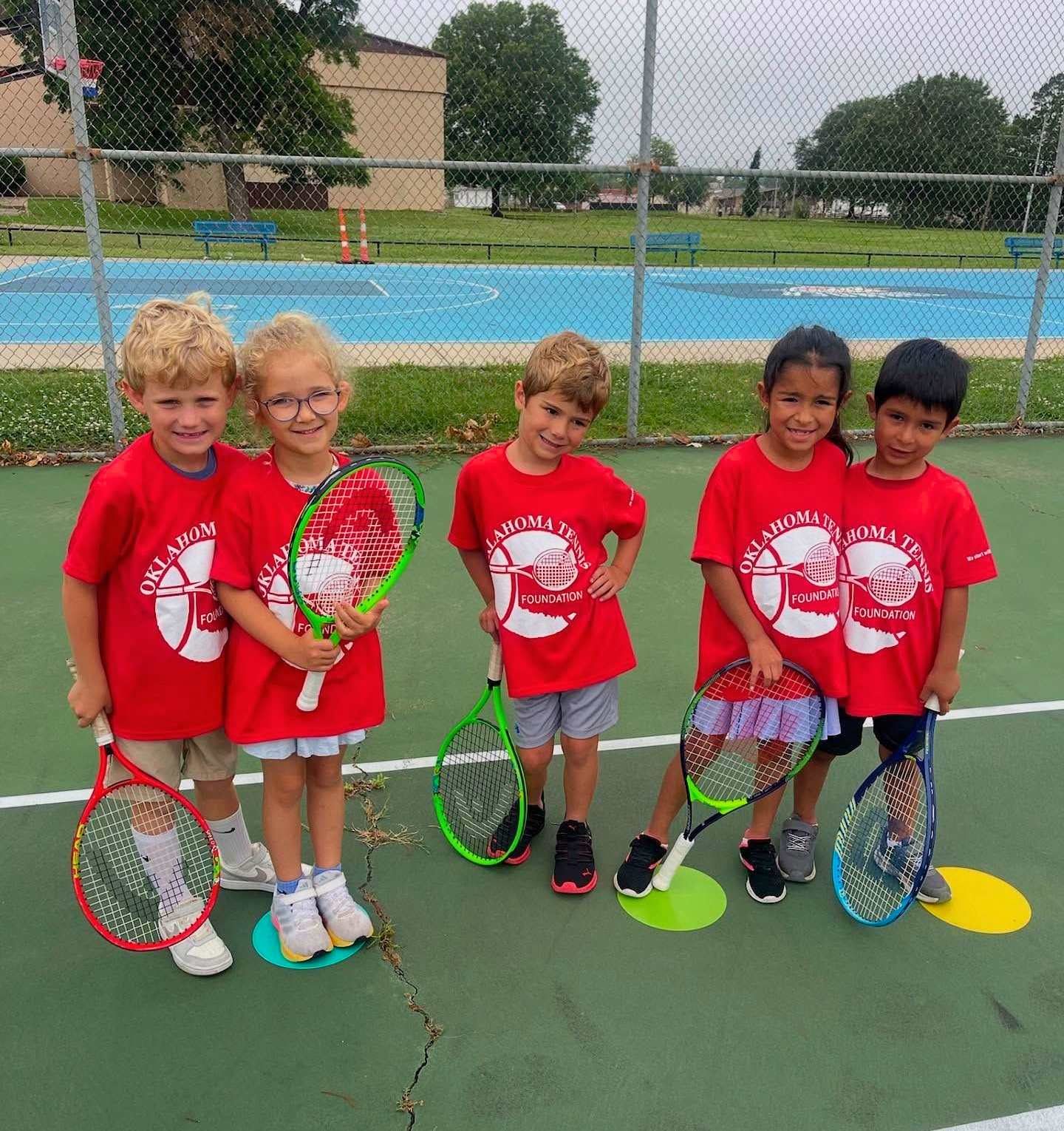 Five children in red tennis shirts stand on a court holding rackets, with a pool and fence behind them.