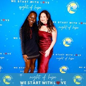 Two women pose at a “Night of Hope” event in front of a blue backdrop with repeated “We Start with Love” text.