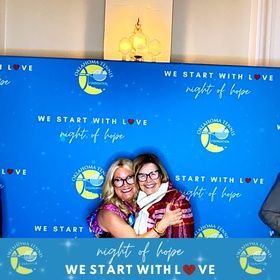 Two women smiling and hugging at a blue “We Start with Love” night of hope event backdrop