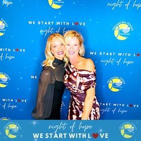 Two women smiling at a blue “We Start With Love” night of hope event backdrop