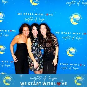 Three women posing at a blue Night of Hope event backdrop with “We Start With Love” text