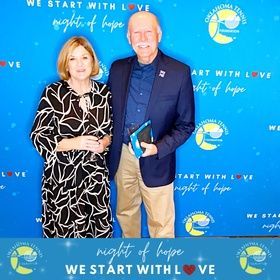Two people posing at a blue “Night of Hope” event backdrop, one holding an award plaque.