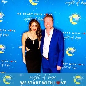 Two people posing at a blue “Night of Hope” event backdrop, smiling and dressed formally.