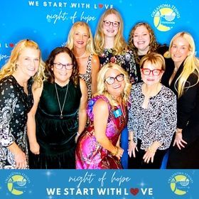 Seven women posing at a Night of Hope event in front of a blue backdrop with hearts and logos.