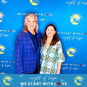 Two people posing in front of a blue “We Start with Love” and “night of hope” event backdrop.