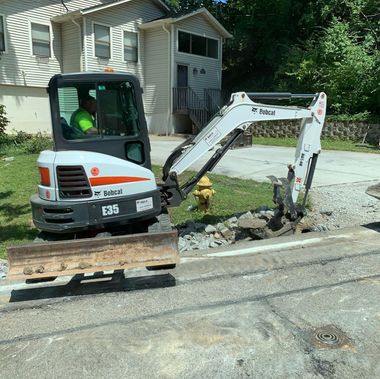 Compact excavator digging up a sidewalk beside a house, with broken concrete and a worker inside the cab.