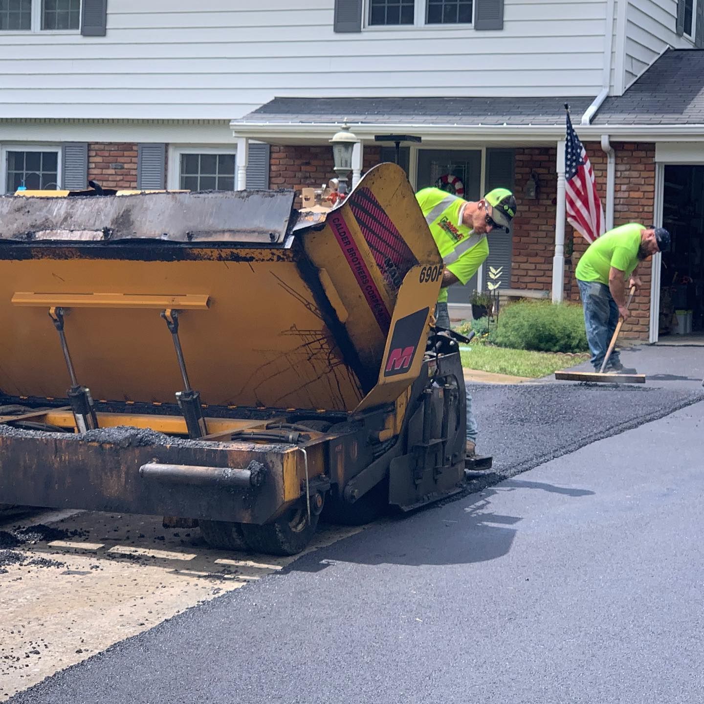 Road paving crew spreading asphalt on a suburban street beside houses and an American flag.
