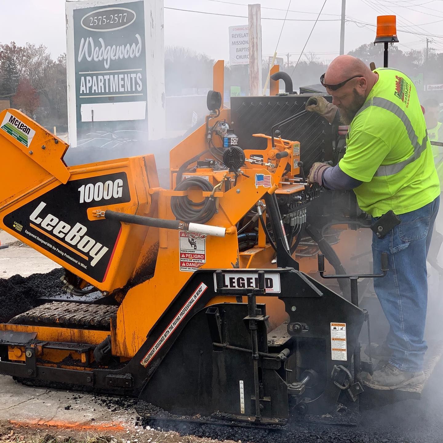 Worker in neon yellow jacket operating an orange LeeBoy paving machine in a snowy street.