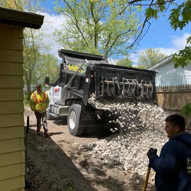 Worker dumps gravel from a truck beside a yellow house while another worker watches.