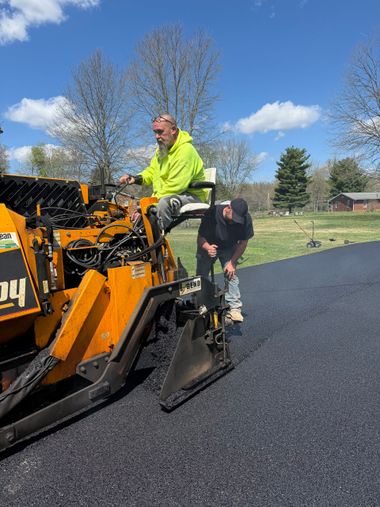 Road crew paving a sidewalk beside a storefront, with an orange paving machine and fresh black asphalt.