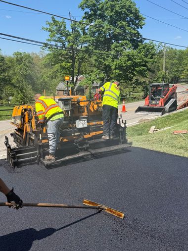 Freshly paved black driveway leading to a beige house with a red roof and trees in the yard