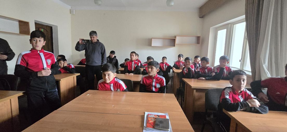 Gruppo di studenti in uniforme rossa seduti in un'aula, con un insegnante in piedi in fondo.