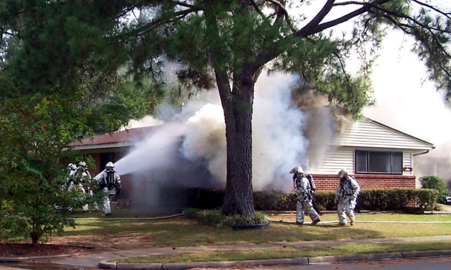 Firefighters in hazmat suits extinguish a house fire, heavy smoke billowing from the building.