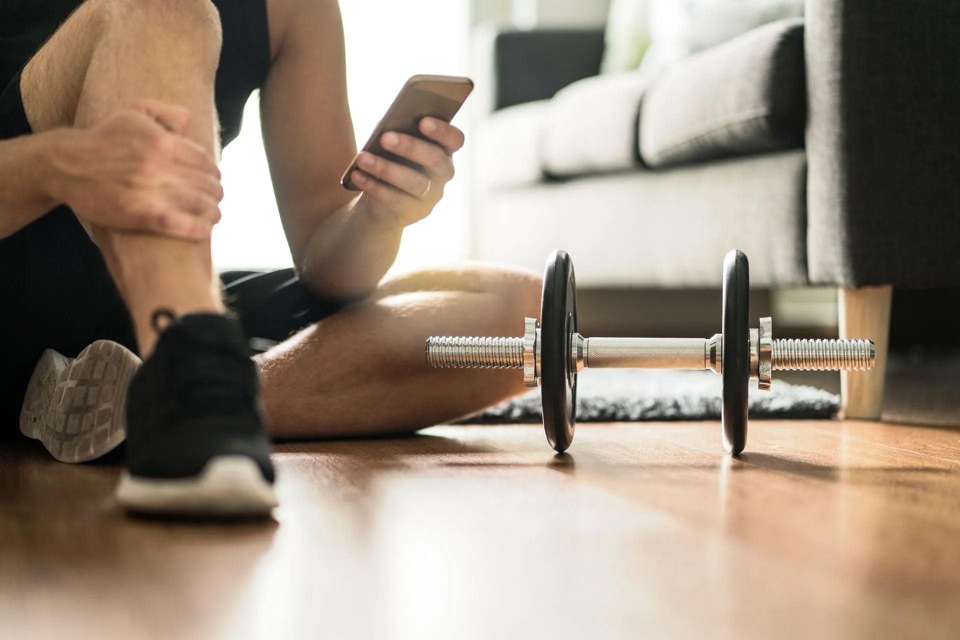 Person stretching on the floor beside a dumbbell, holding a phone in a bright living room