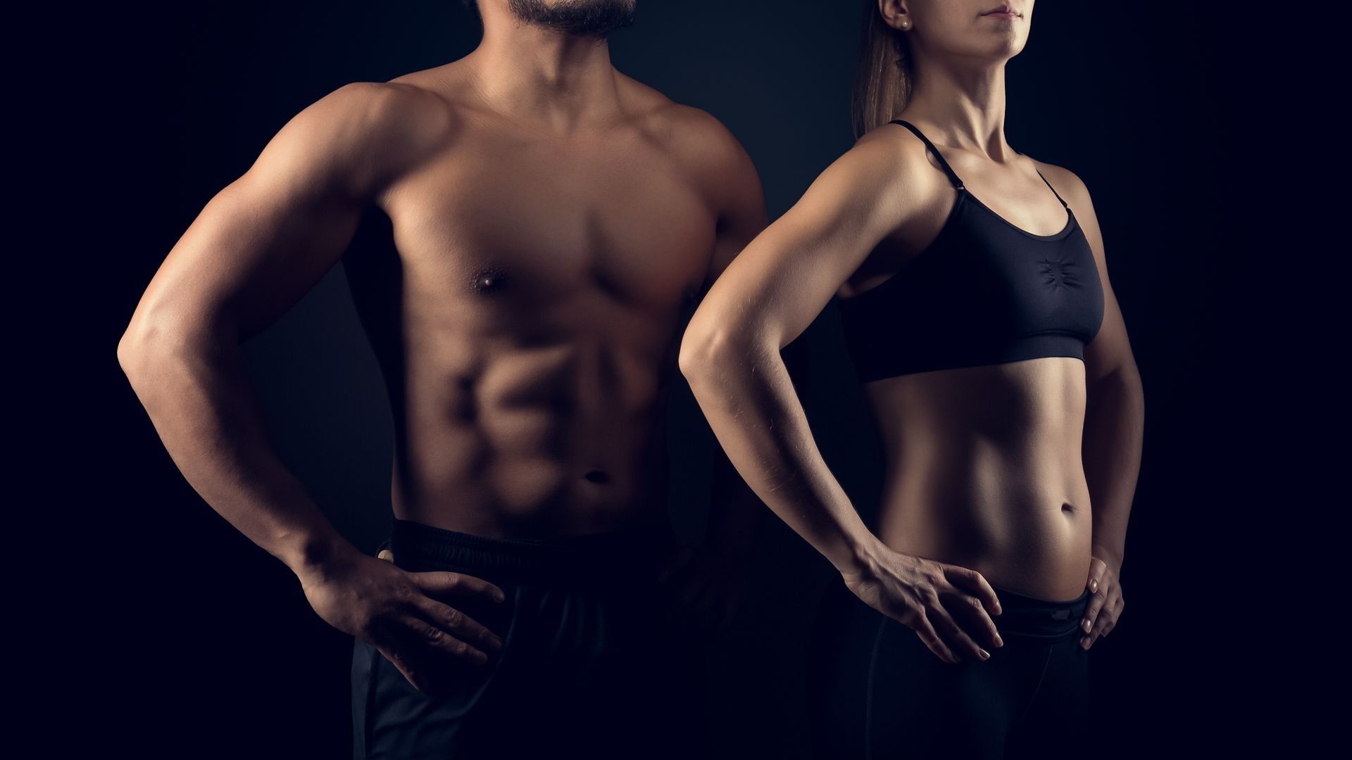 Two fit adults in dark workout clothes standing side by side against a dark background, hands on hips.