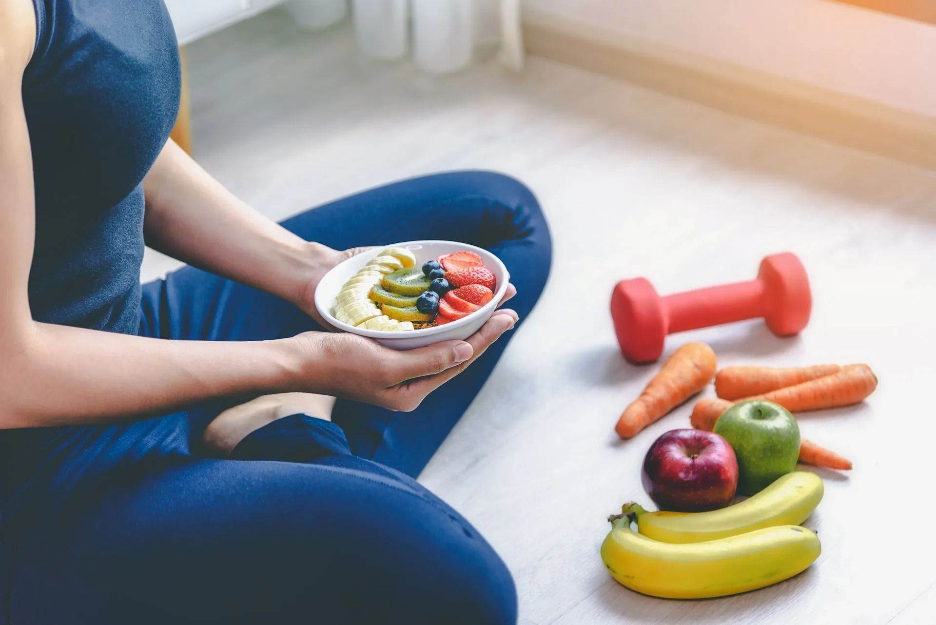 Person sitting with a healthy snack bowl beside fruits, vegetables, and red dumbbells on the floor.