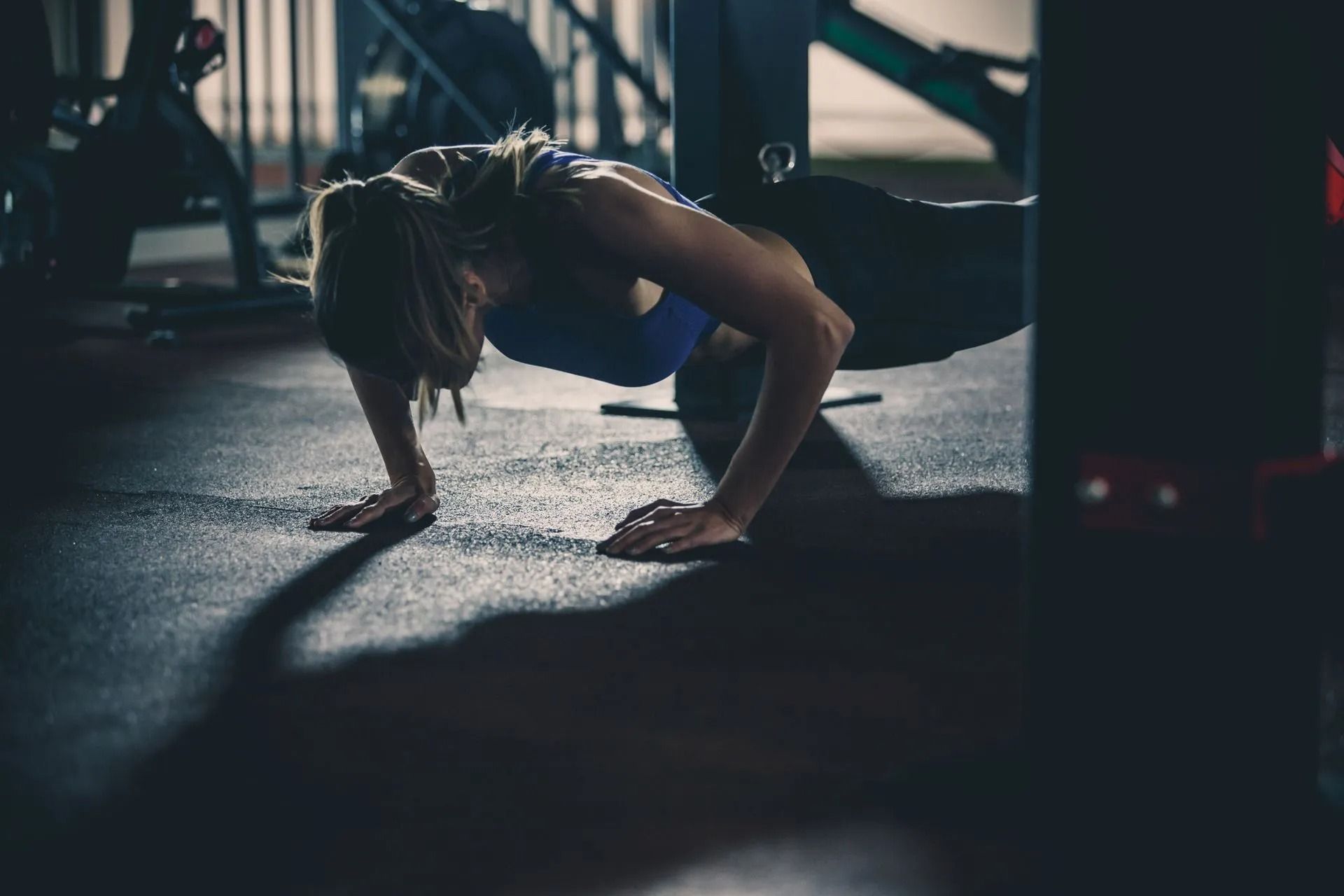 Person doing a push-up in a dim gym, lit by side light on a rubber floor.