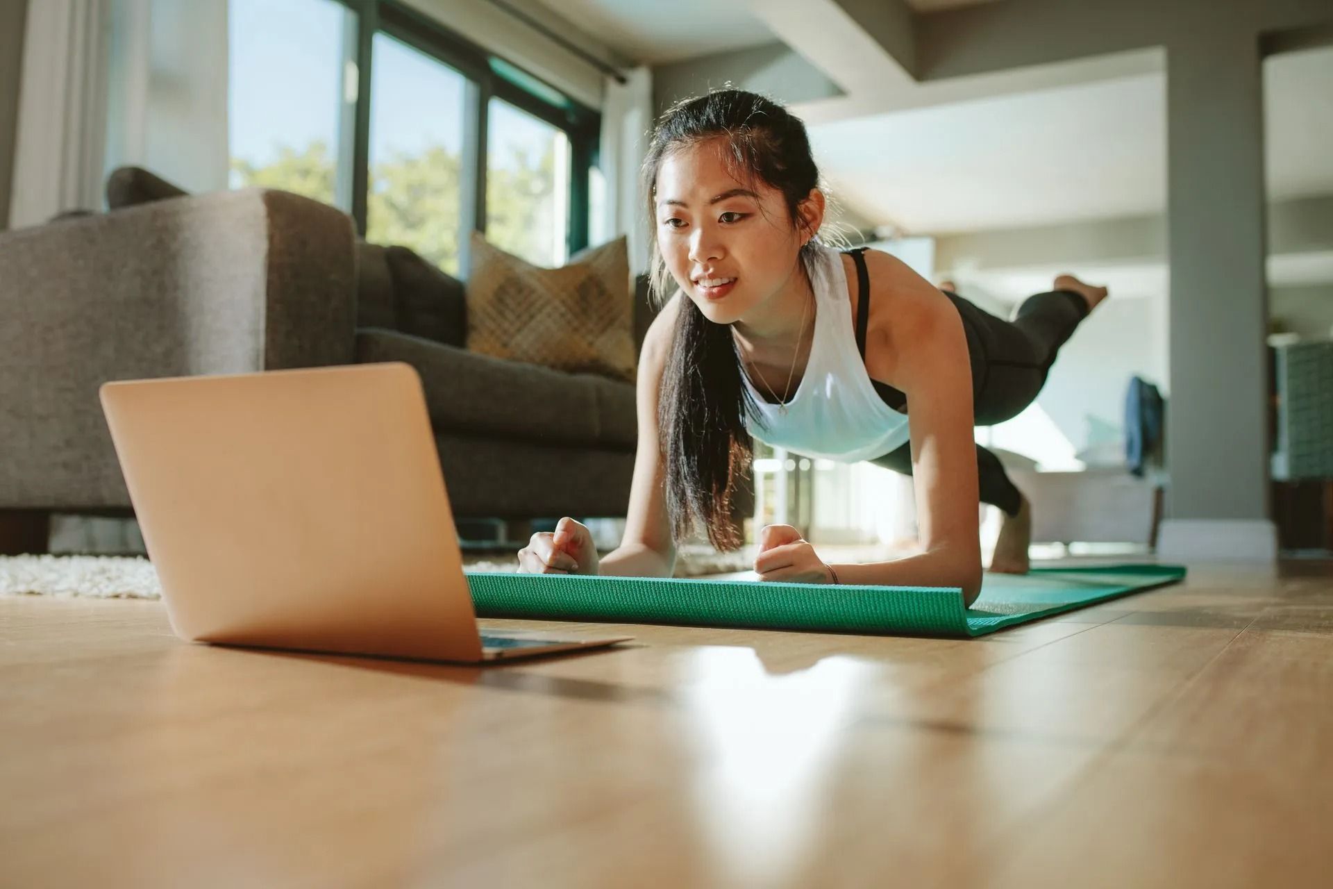 Woman doing a yoga plank on a mat beside a laptop in a bright living room