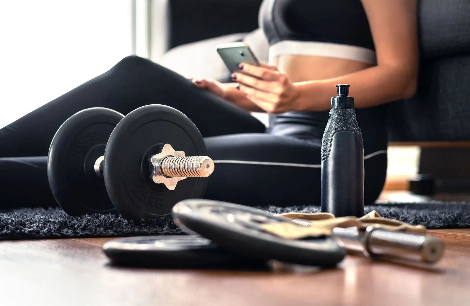 Person relaxing on a mat with a phone beside black dumbbells and a water bottle