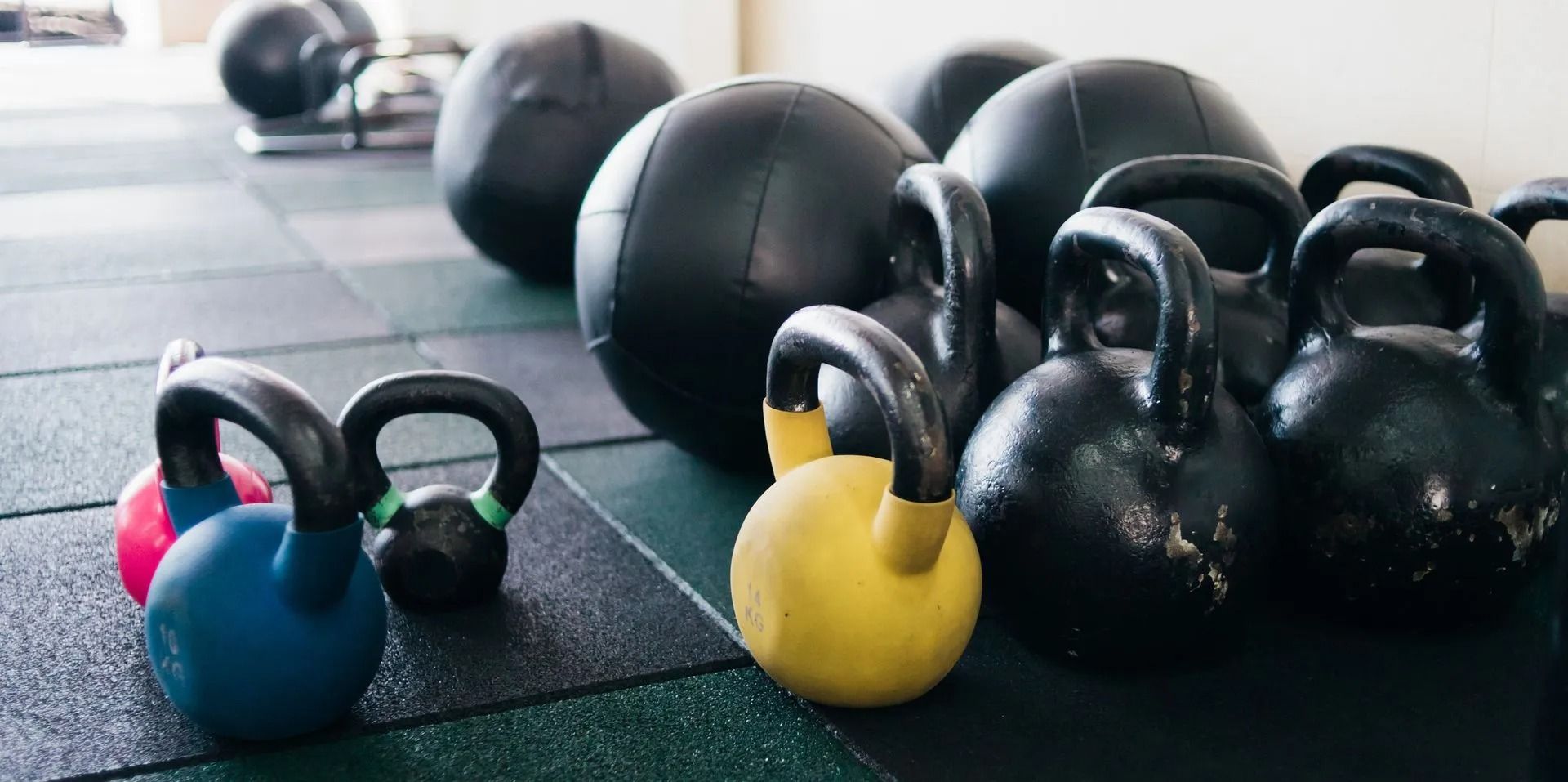 Kettlebells and medicine balls on a gym floor, including blue, yellow, and black weights.