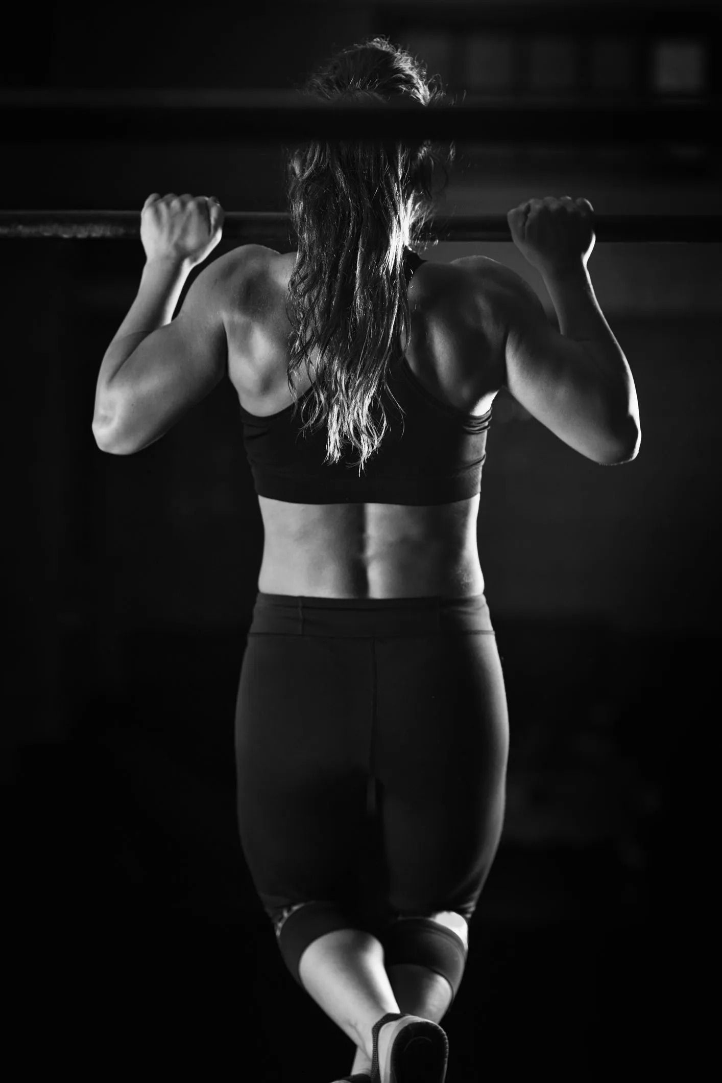 Woman doing pull-ups on a bar in a dark gym, seen from behind in black athletic wear