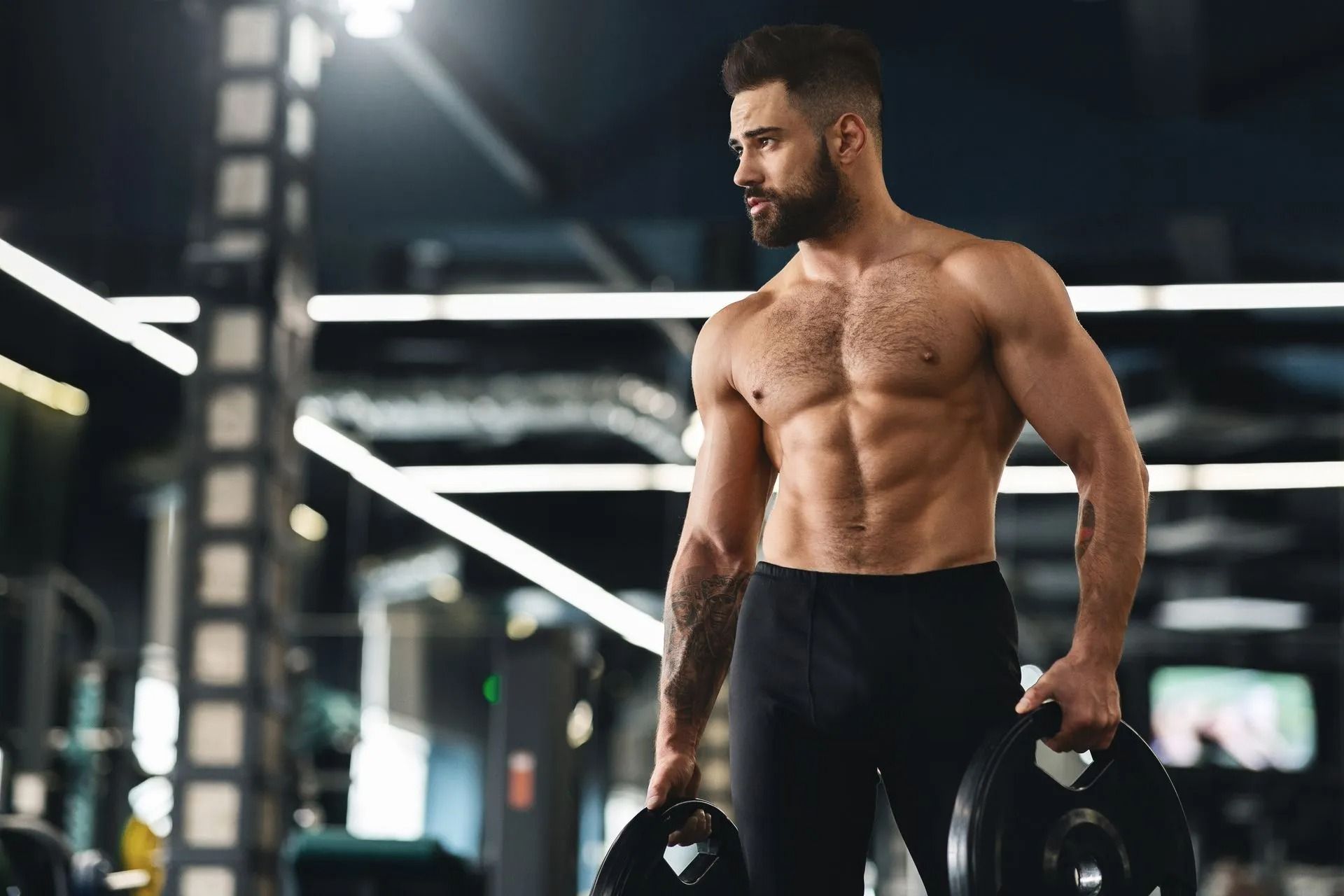 Muscular man holding dumbbells in a gym, shirtless and wearing black pants