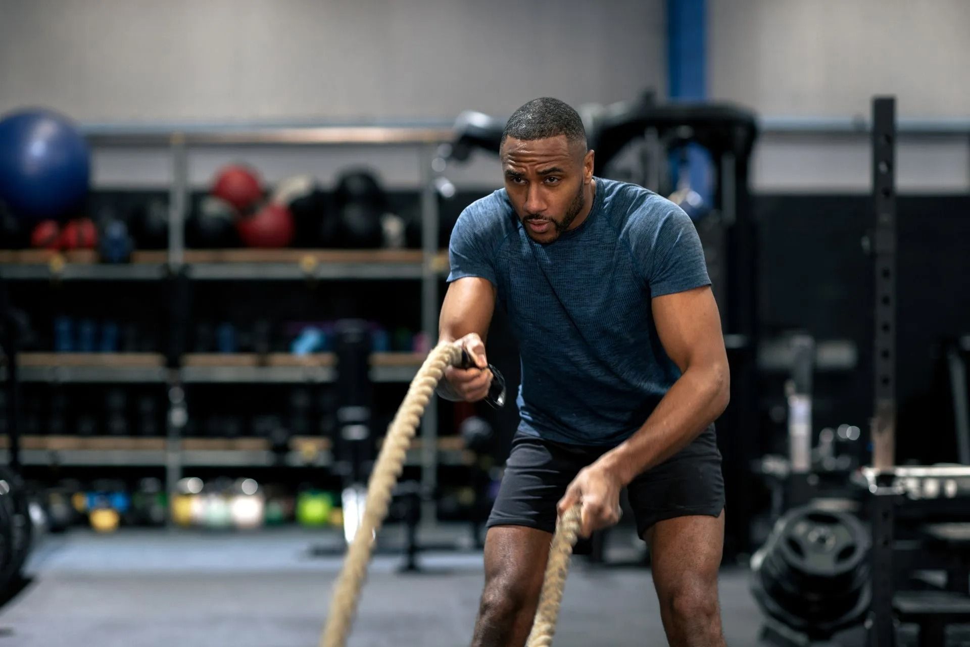Man battling battle ropes in a gym, focused and leaning forward in a dark workout shirt.