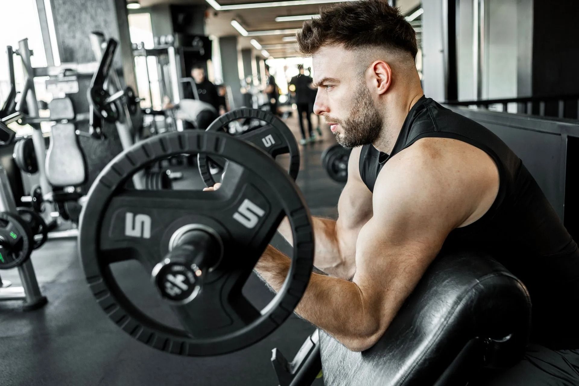 Man lifting a barbell in a gym, seated at a preacher curl bench.